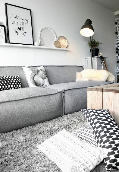 Ο γκρι καναπές σε γεωμετρικό ογκώδες σχήμα συνδυάζεται με μαύρο φωτιστικό, ξύλινο coffee table και πλούσιο χαλί στο ίδιο χρώμα.